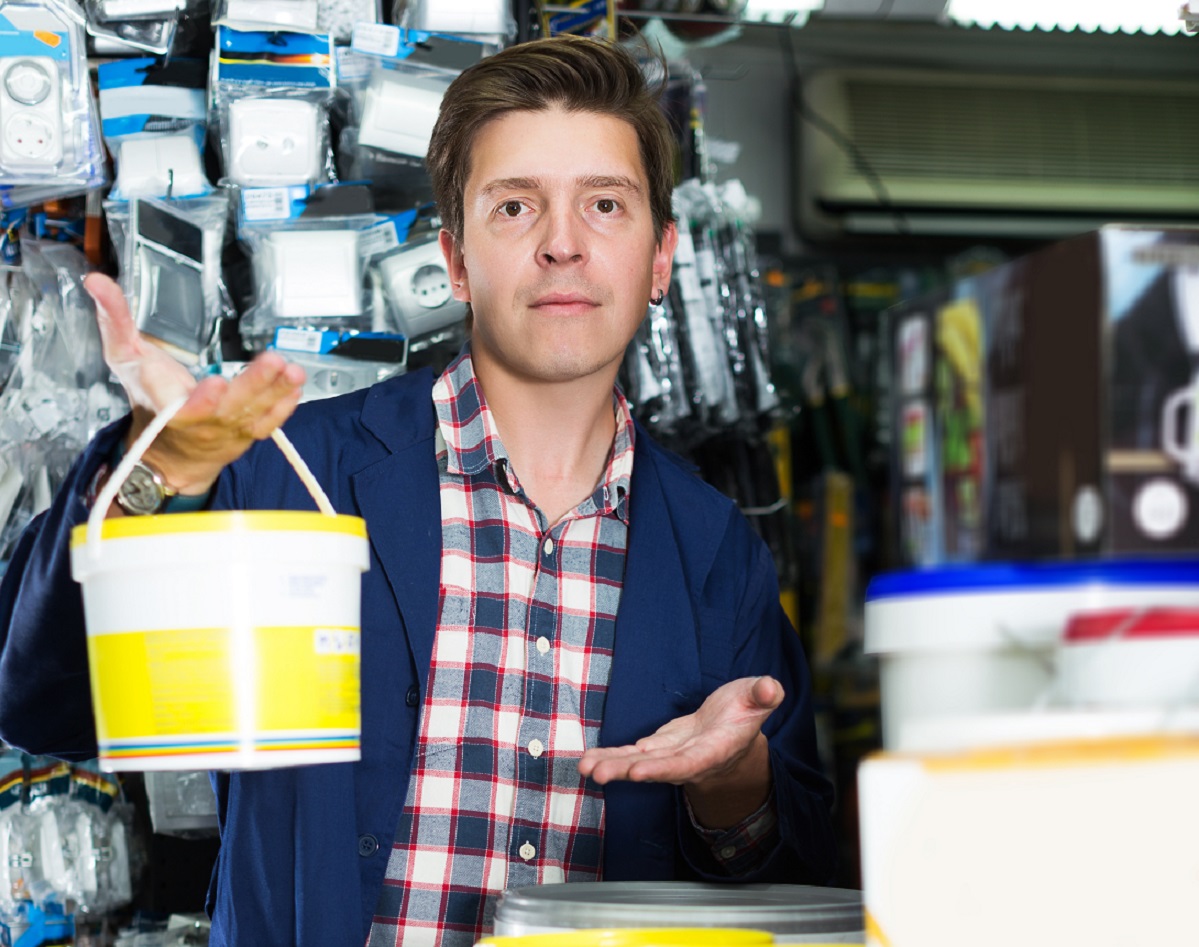 Young cheerful worker selling tins of paint in hardware shop