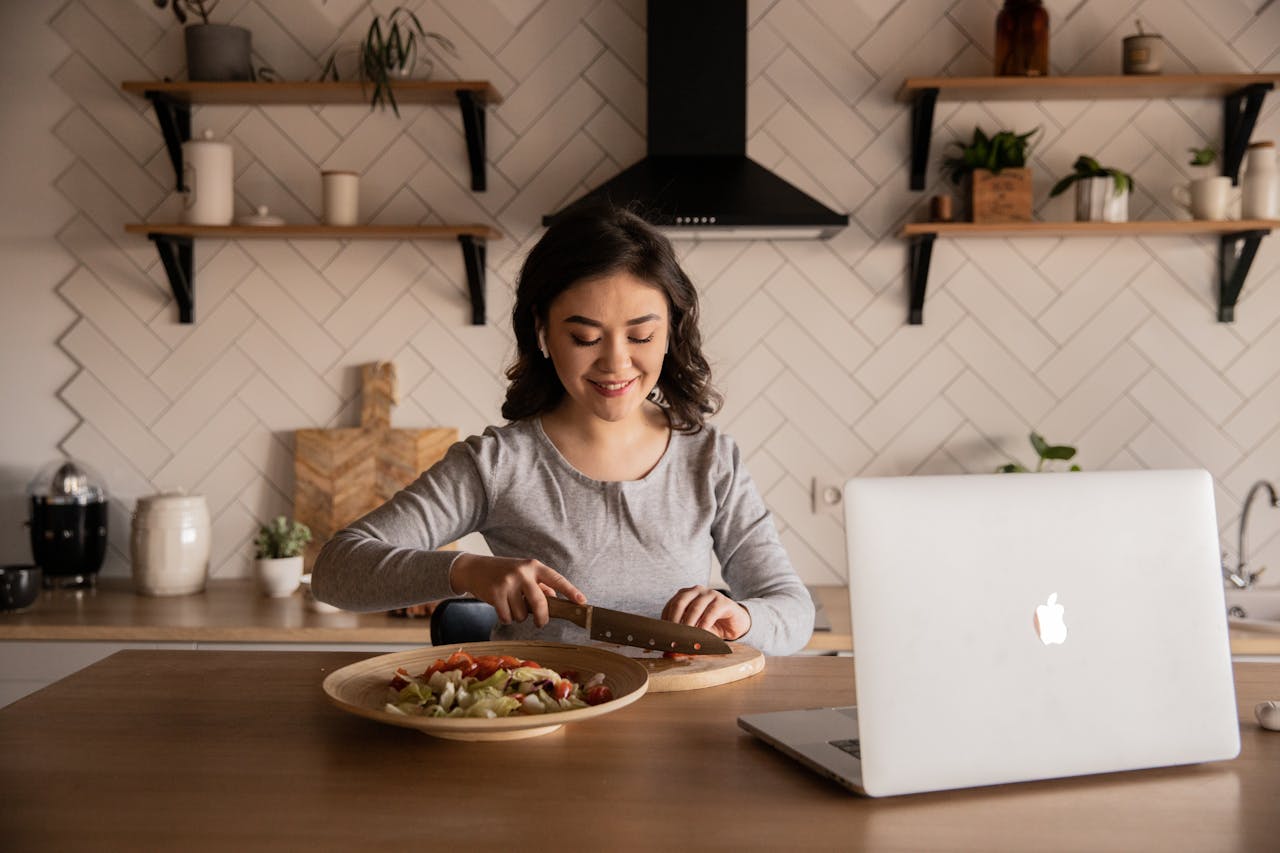 Young woman preparing vegetable salad in front of open laptop placed on a wooden table