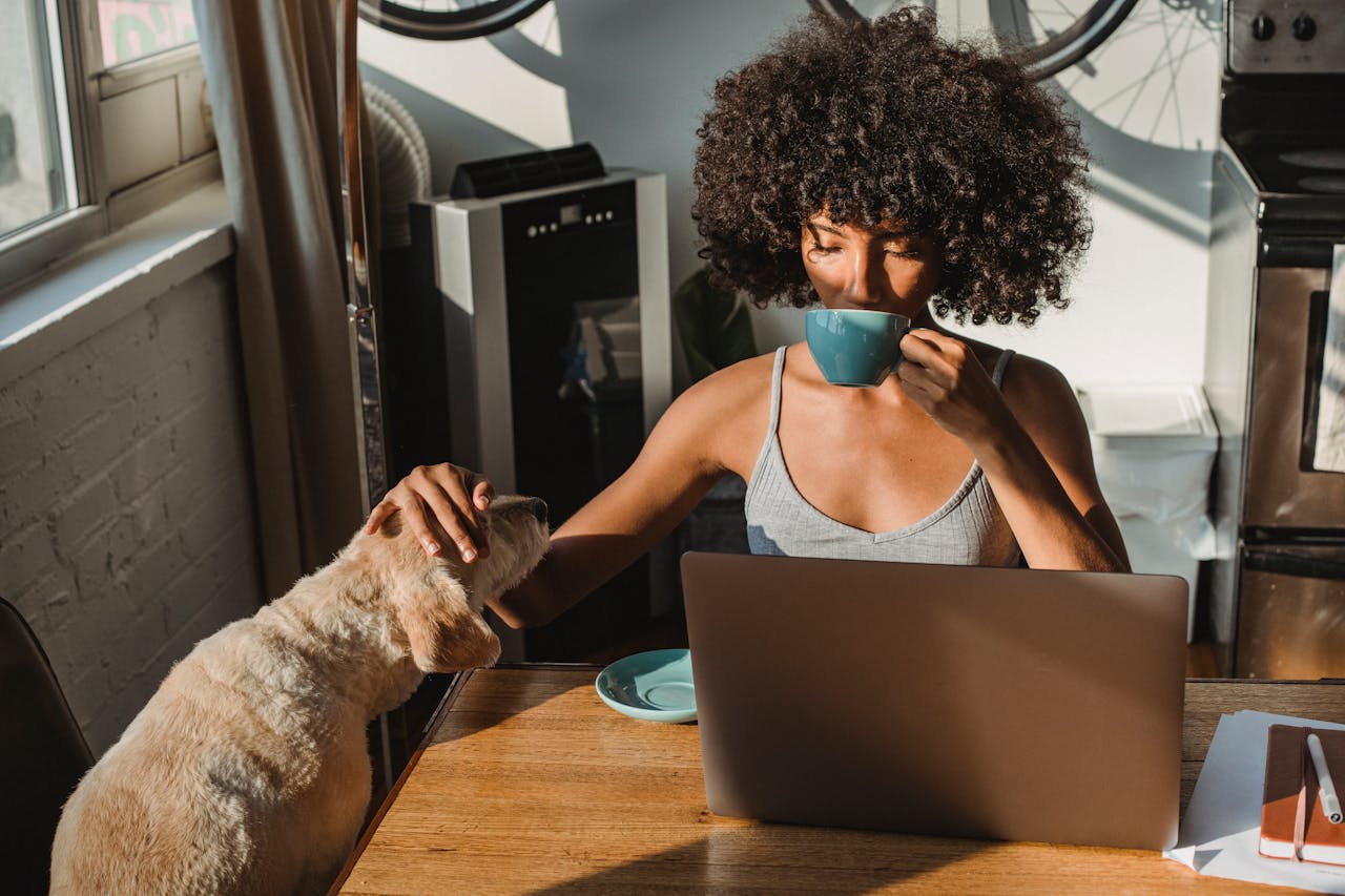 Young female freelancer sitting on a wooden table, petting a dog, using laptop and drinking coffee