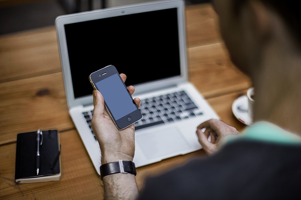 A man holding an iPhone while sitting at a wooden desk with a laptop