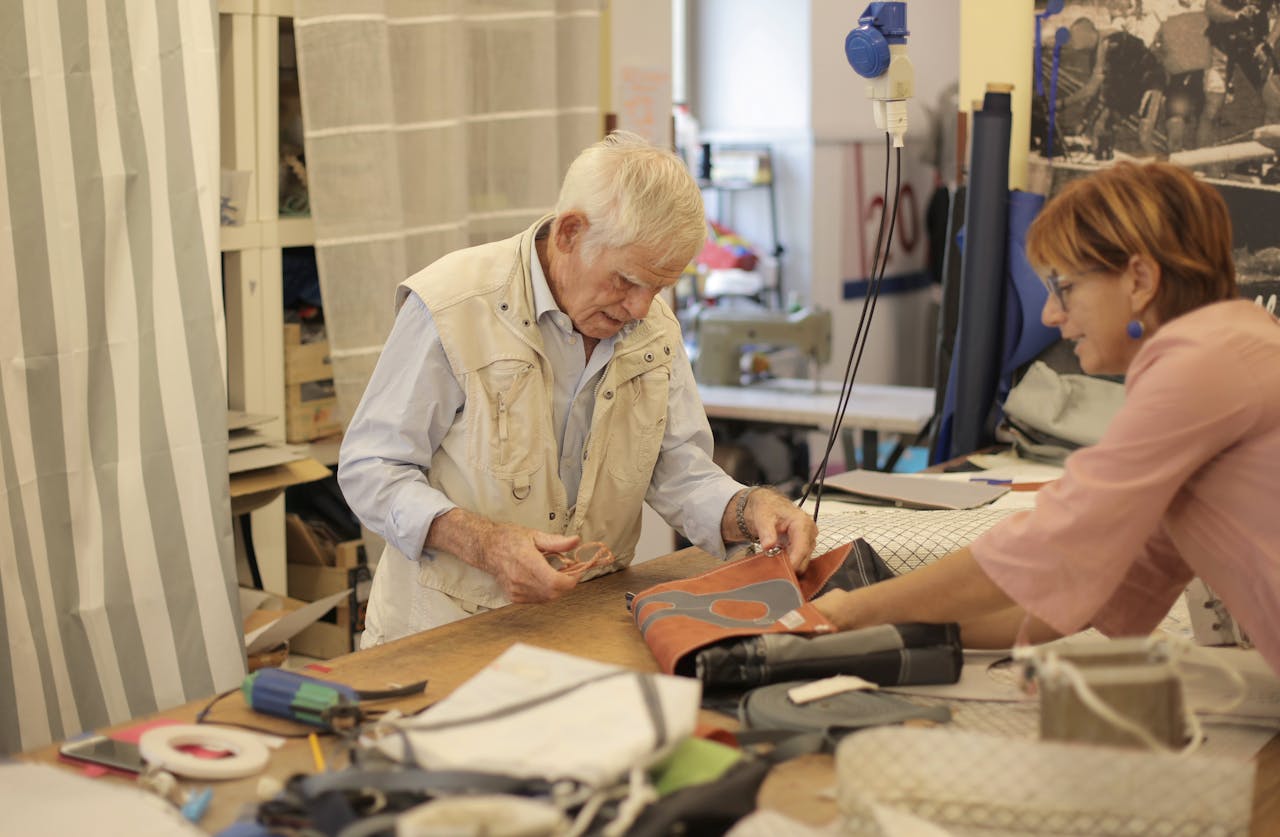 Aged craftsman making leather bag in workshop with assistant