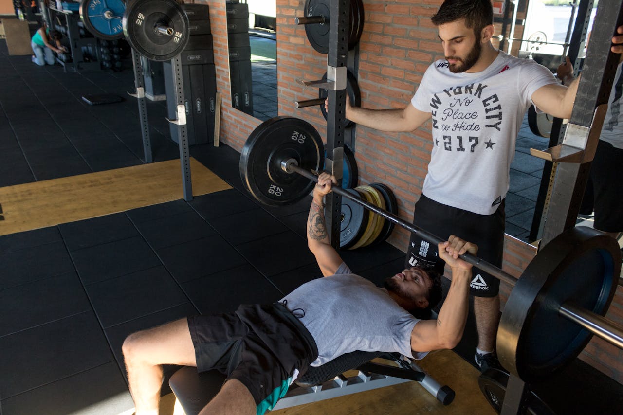 Photo of a man lying while doing barbell in a gym next to his personal trainer