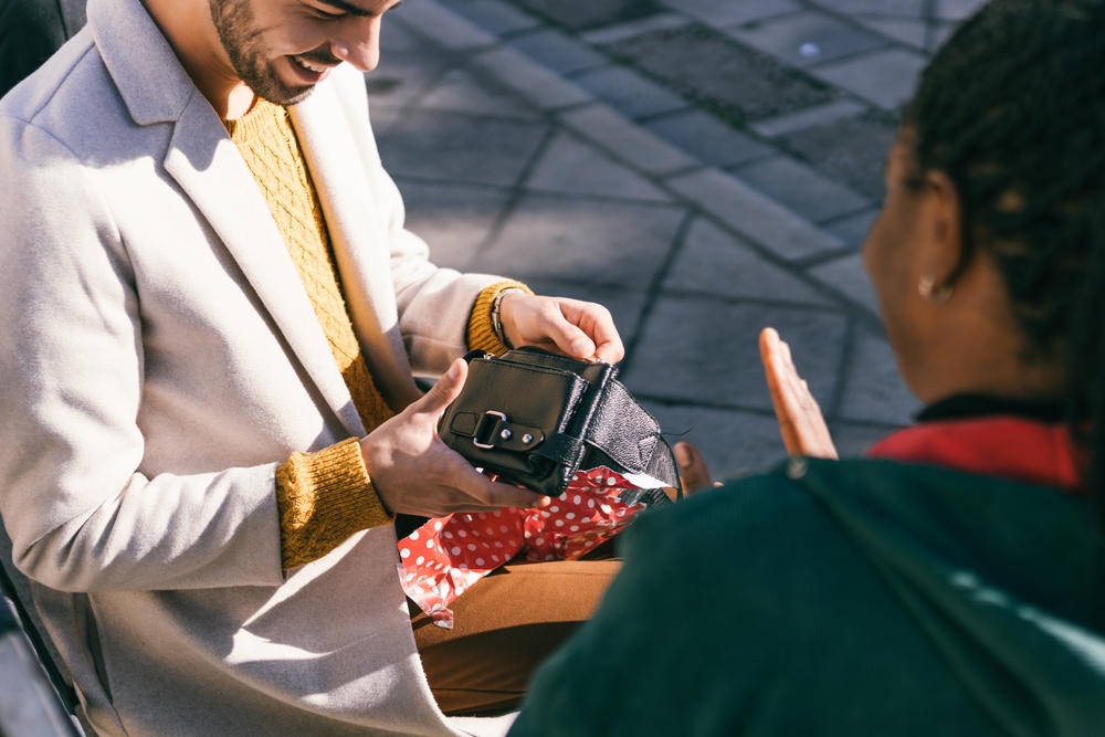 Man opening a bag with a leather purse inside sitting on a bench next to a friend