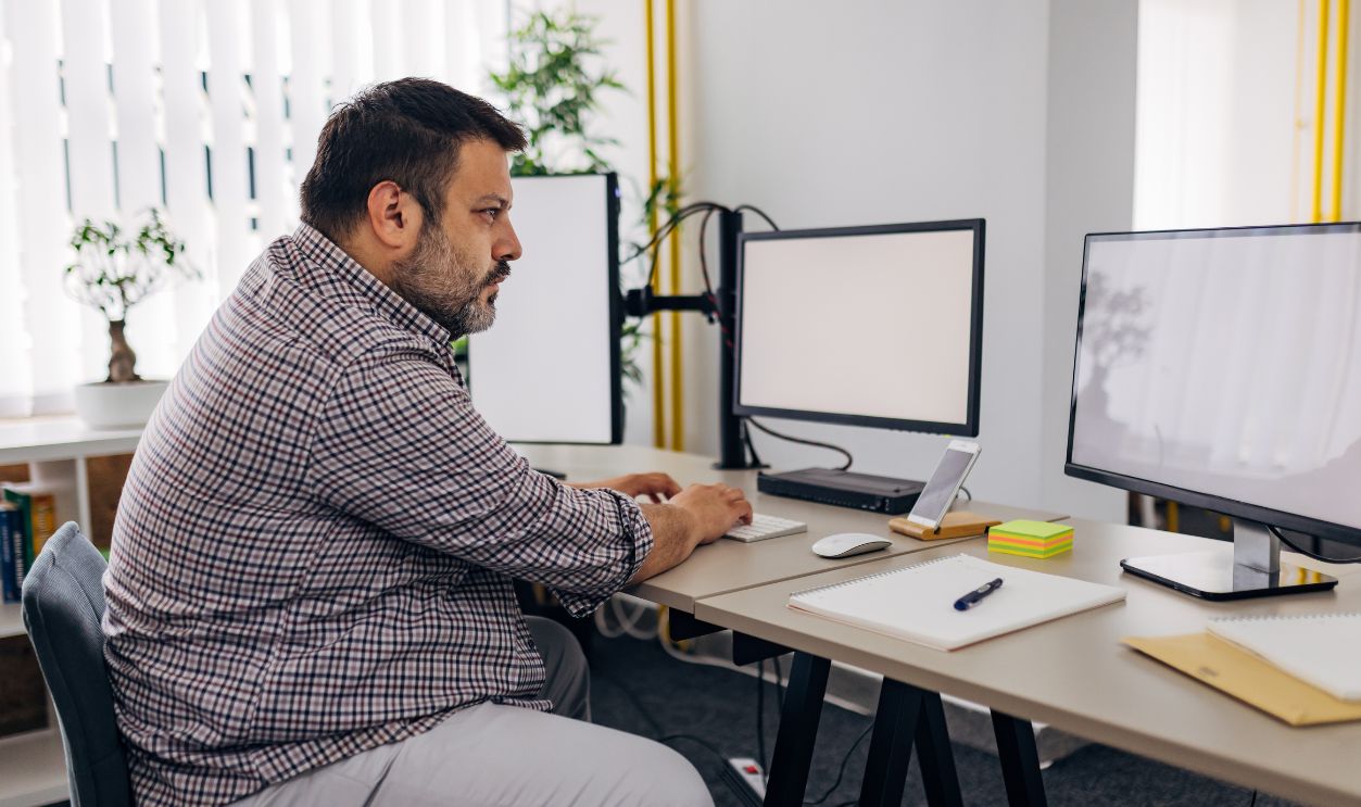 Gettyimages - 1498126281, Male software developer working with computers and data systems in office - stock photo A male IT professional is sitting at a desk in the office and working on a computer