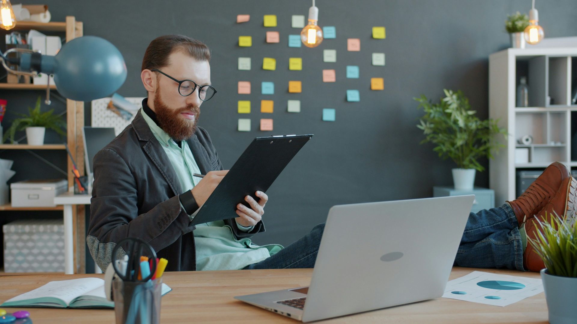 Man with beard working at desk with laptop and papers.