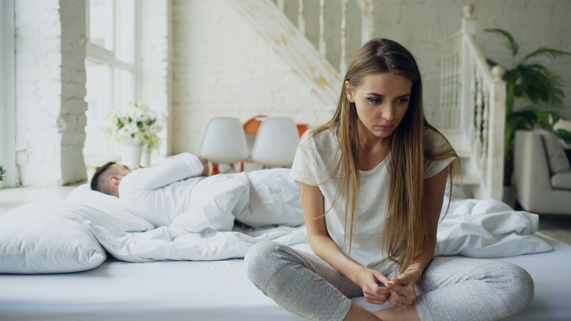 Woman sitting on bed, man sleeping behind her.