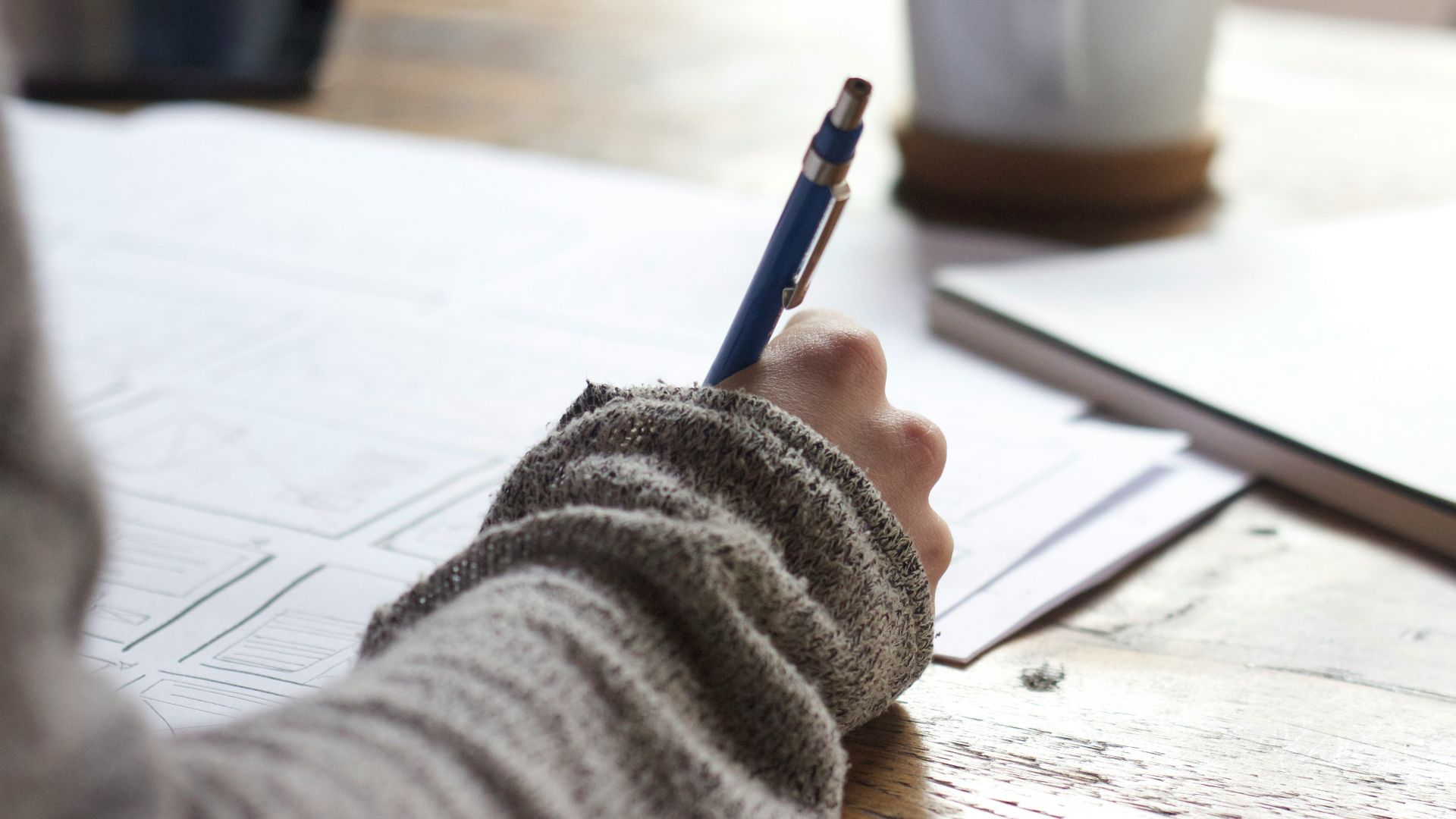 person writing on brown wooden table near white ceramic mug