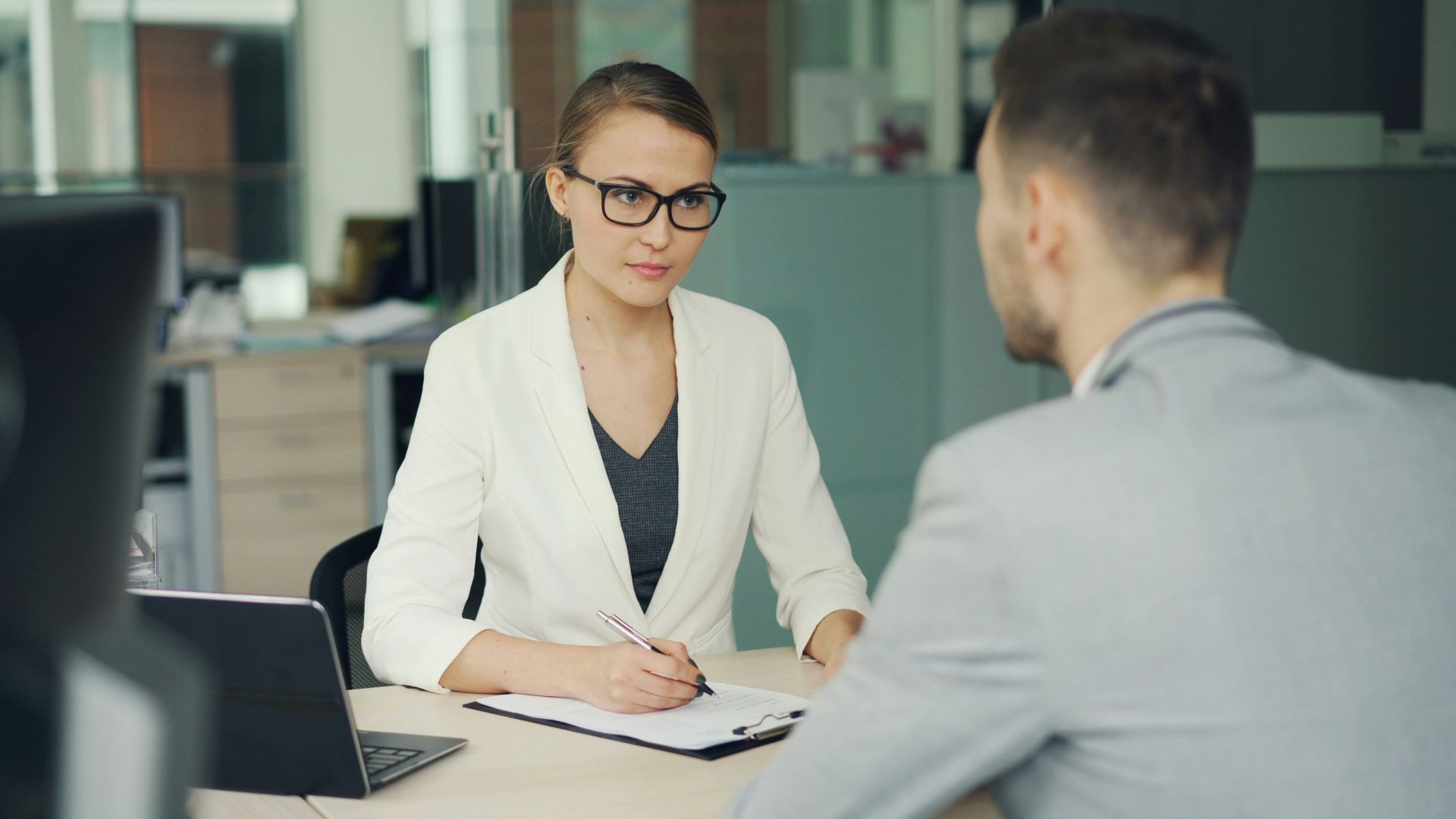 Woman in glasses interviews man at office desk.