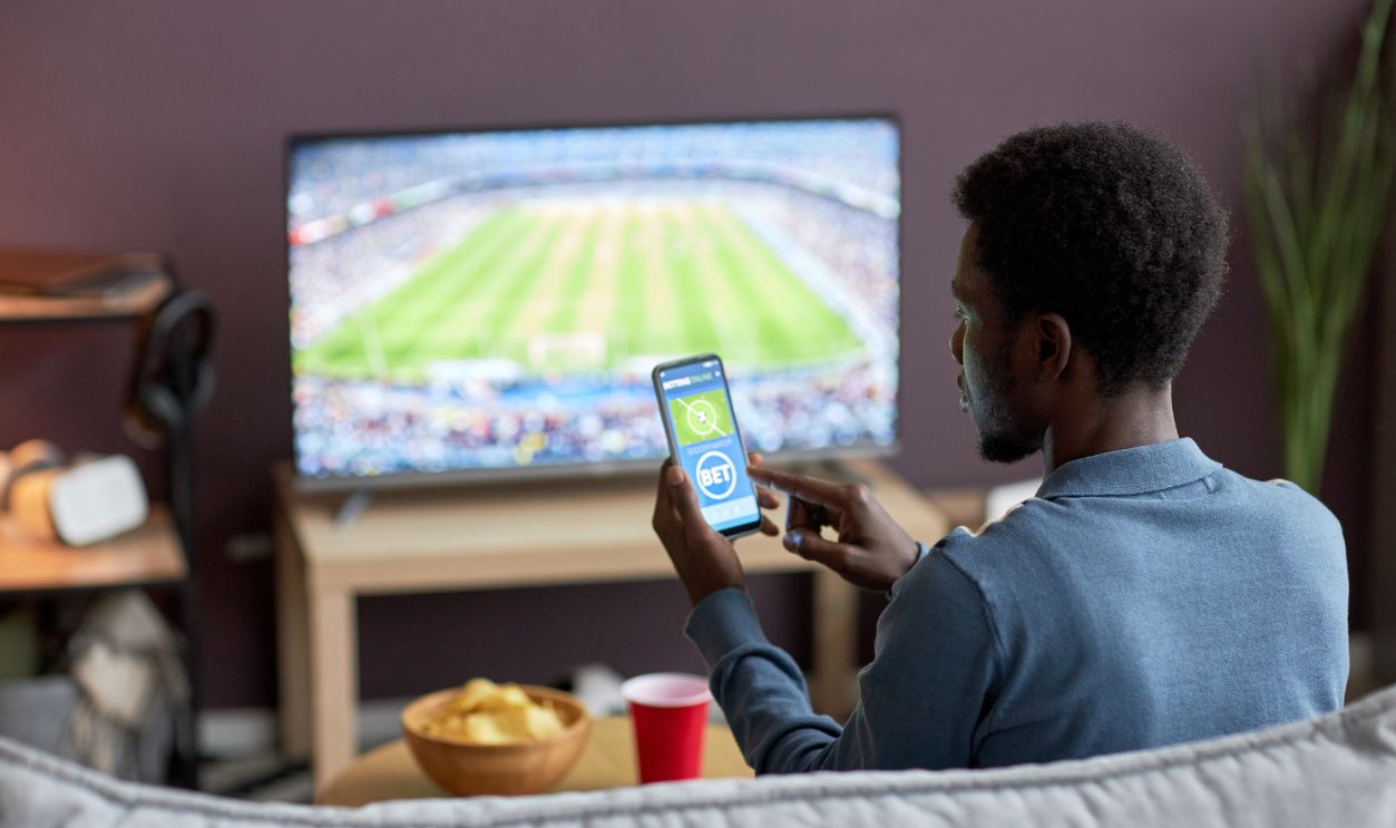 Back view of man holding smartphone with sports bets app on screen while watching football match at home, copy space