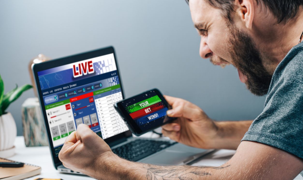 Young attractive bearded man showing sincere excitement about his favorite team victory. Guy being happy winning a bet in online sport gambling application on his mobile phone.