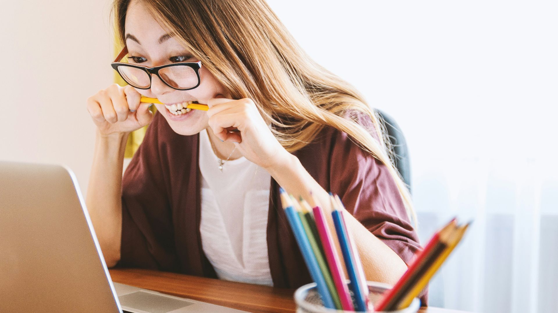 woman biting pencil while sitting on chair in front of computer during daytime