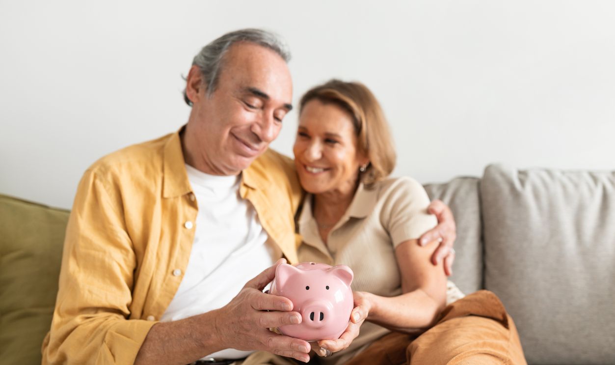 Happy european senior couple holding pink piggybank in hands and embracing, sitting on sofa in living room interior, free space. Retirement savings concept