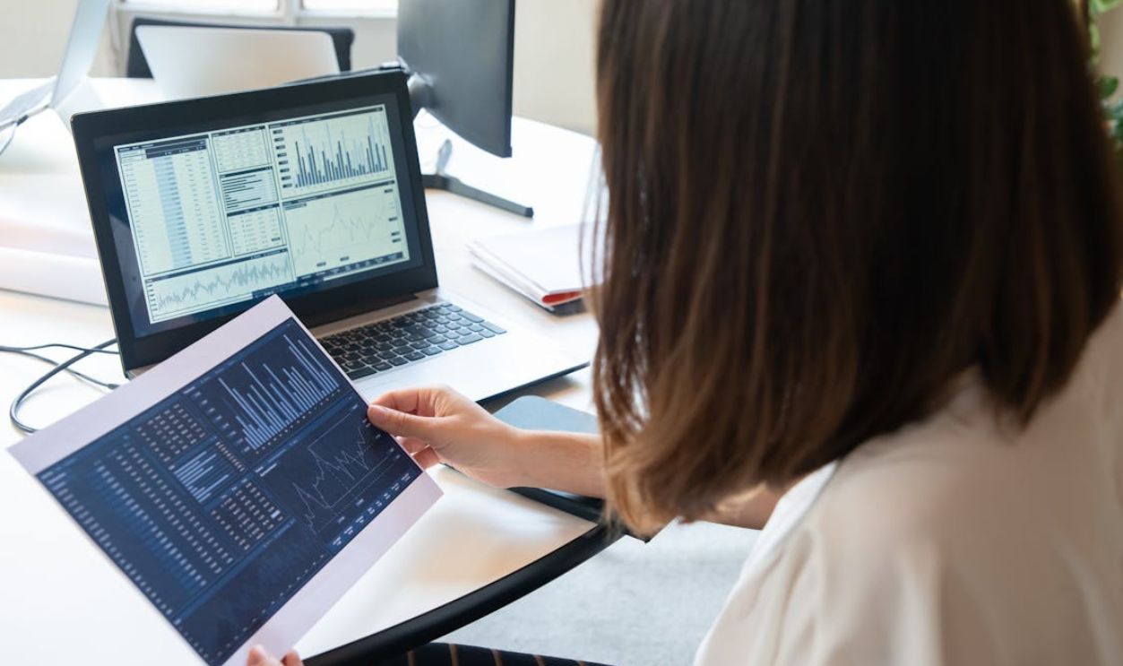 Photo of a Woman Holding a Paper with Charts Near Her Laptop