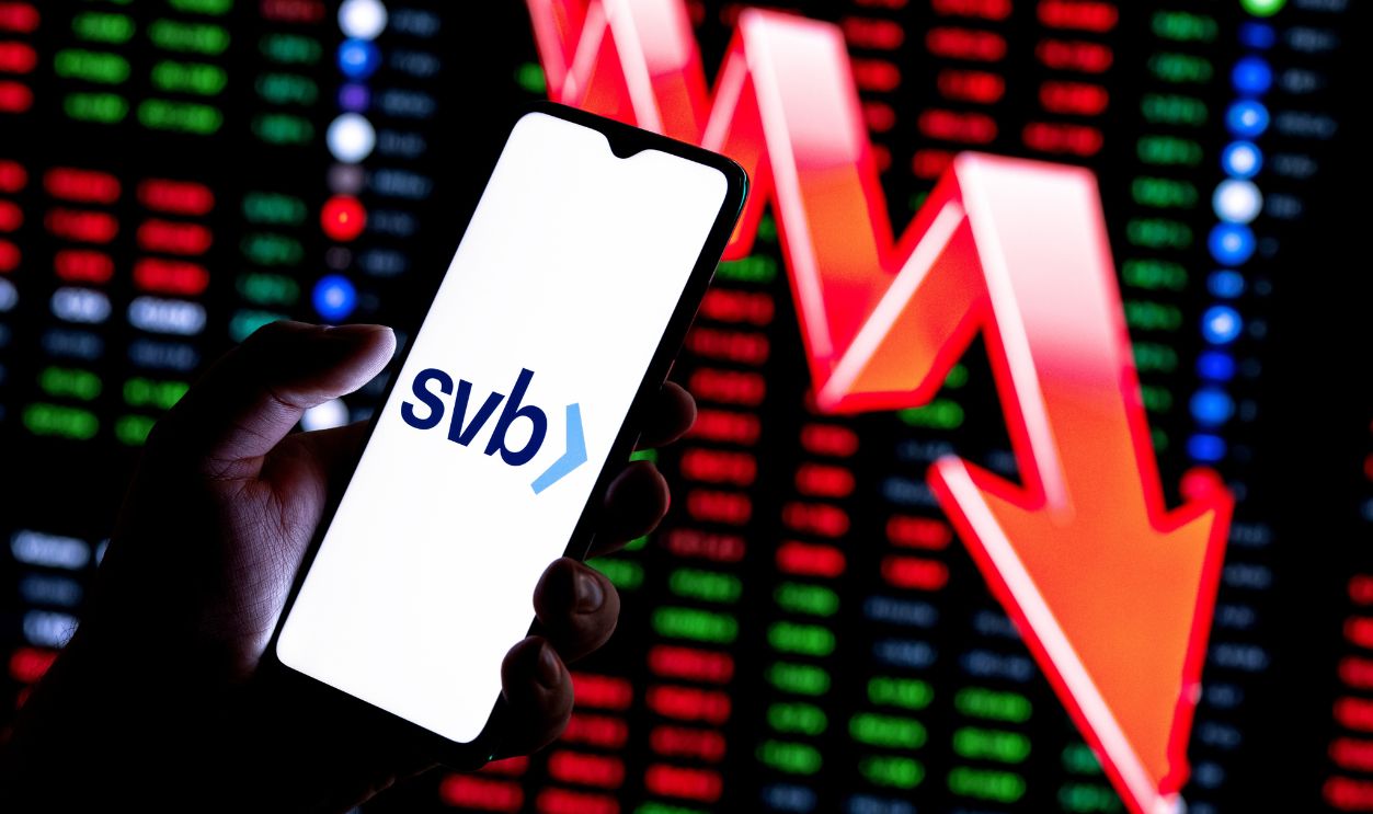 Kazan, Russia - Mar 13, 2023: Smartphone with Silicon Valley Bank logo in hand on background of large red arrow pointing down. Falling stock price.