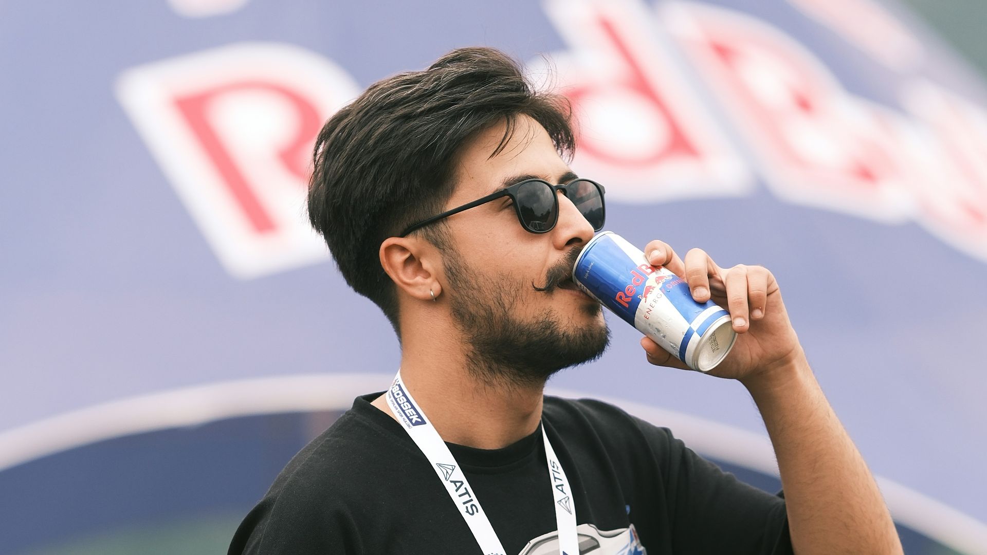 A man drinking from a bottle while standing in front of a red bull tent