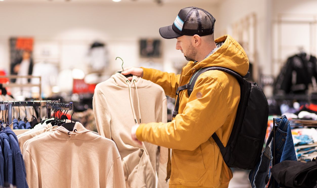 Bearded Caucasian young man wearing backpack chooses clothes in store. Concept of shopping and discounts.
