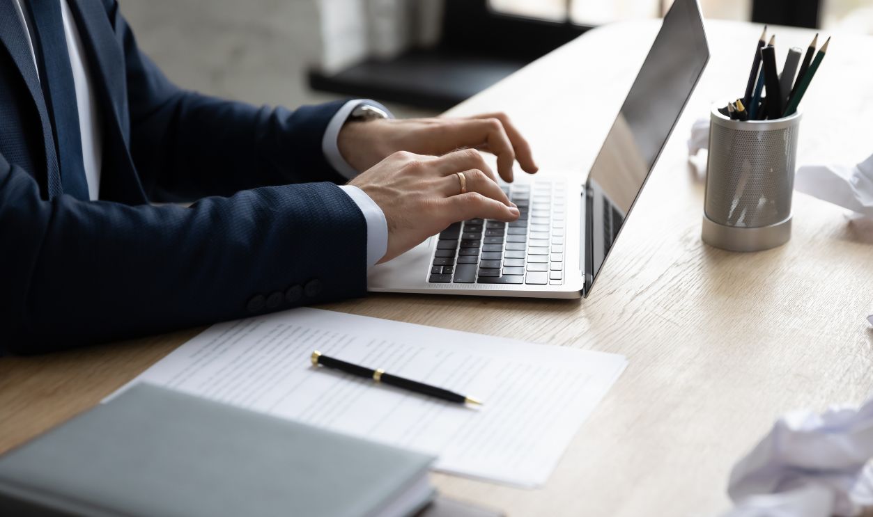 Businessman using laptop among crumpled paper drafts messy in office. Professional typing important document on keyboard, writing article or report, working at computer. Close up of hands