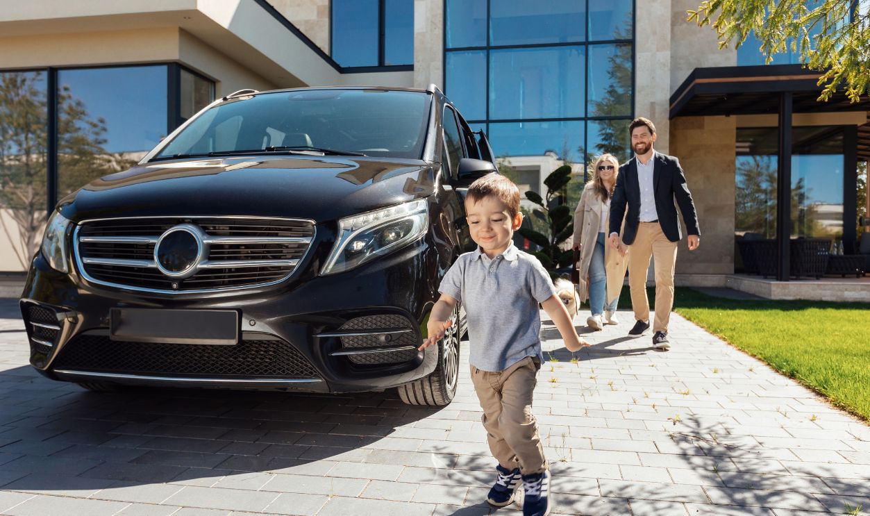 Little boy is running on yard of house near luxury car with parents on background