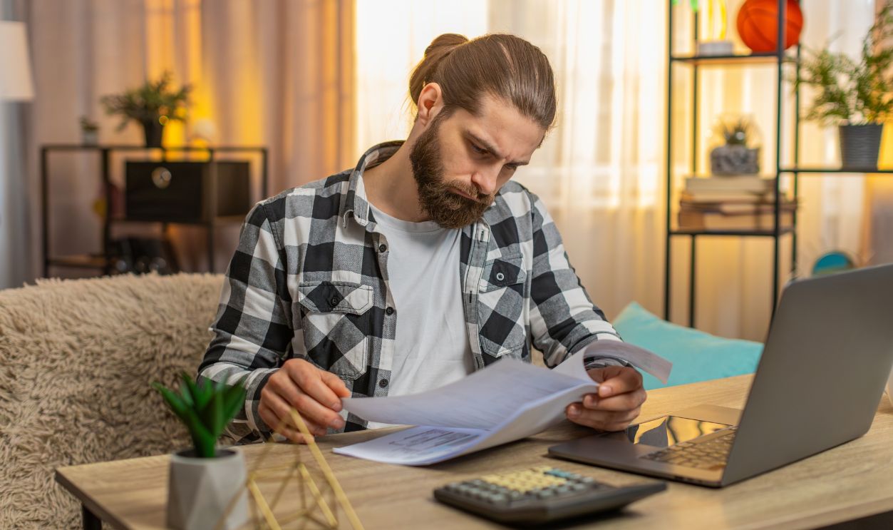 Young serious man checks papers at home table reading fine print new agreement with deep concern studying analyzes contract with a bank, clutches head at complex conditions clauses feeling pressured.