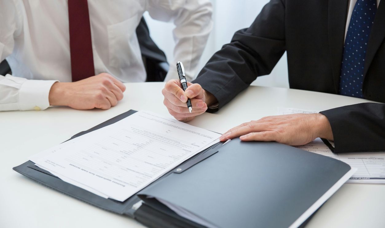 A Person in Black Suit Holding a Pen Near the Documents on the Table