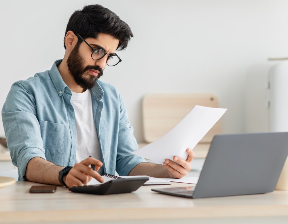 Young man using calculator and laptop computer, sitting at kitchen
