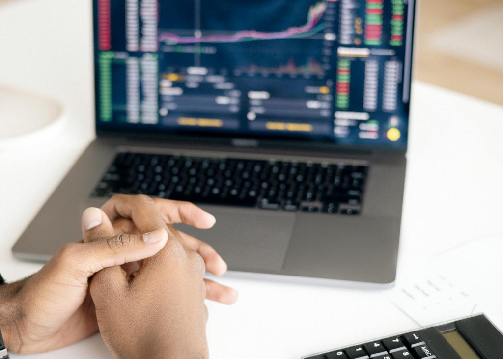 The image shows a close-up of a pair of hands clasped together in front of a laptop displaying financial data