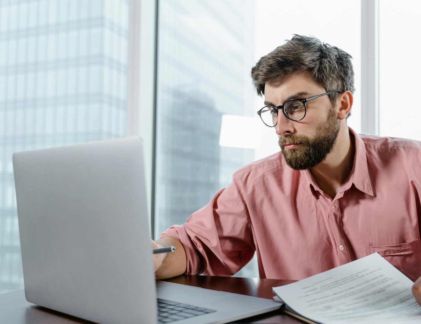 The image shows a man with short brown hair, a beard, and round-framed glasses sitting at a desk and working on a laptop