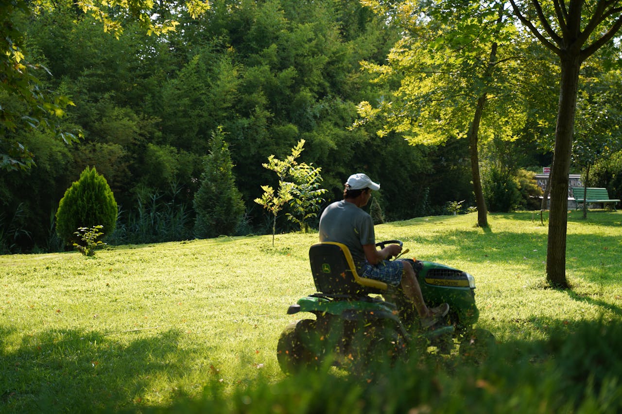 The image shows a person operating a green and yellow John Deere riding lawn mower on a sunny day