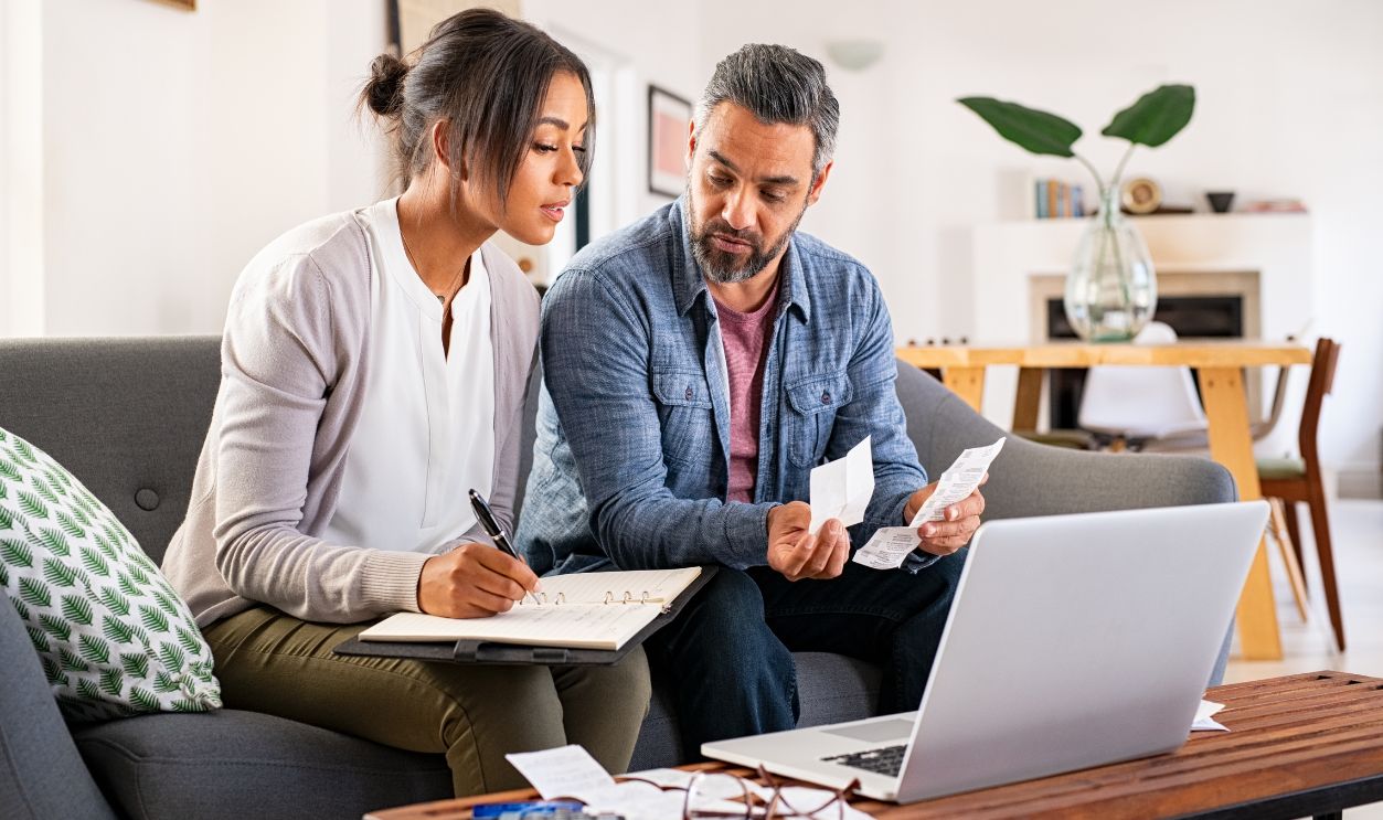 Mature couple calculating bills at home using laptop and calculator. Multiethnic couple working on computer while calculating finances sitting on couch. Mature indian man with african american woman at home analyzing their finance with documents.