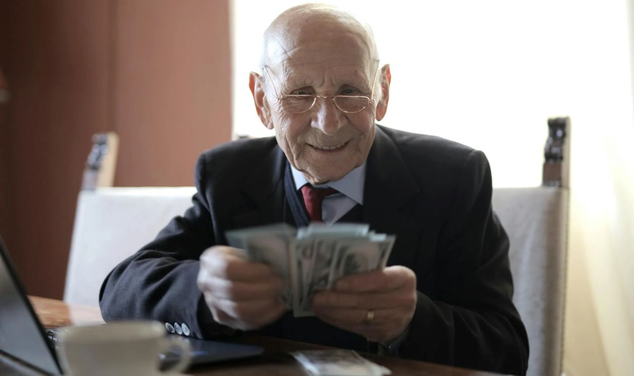 Happy senior businessman counting money while sitting at table with laptop