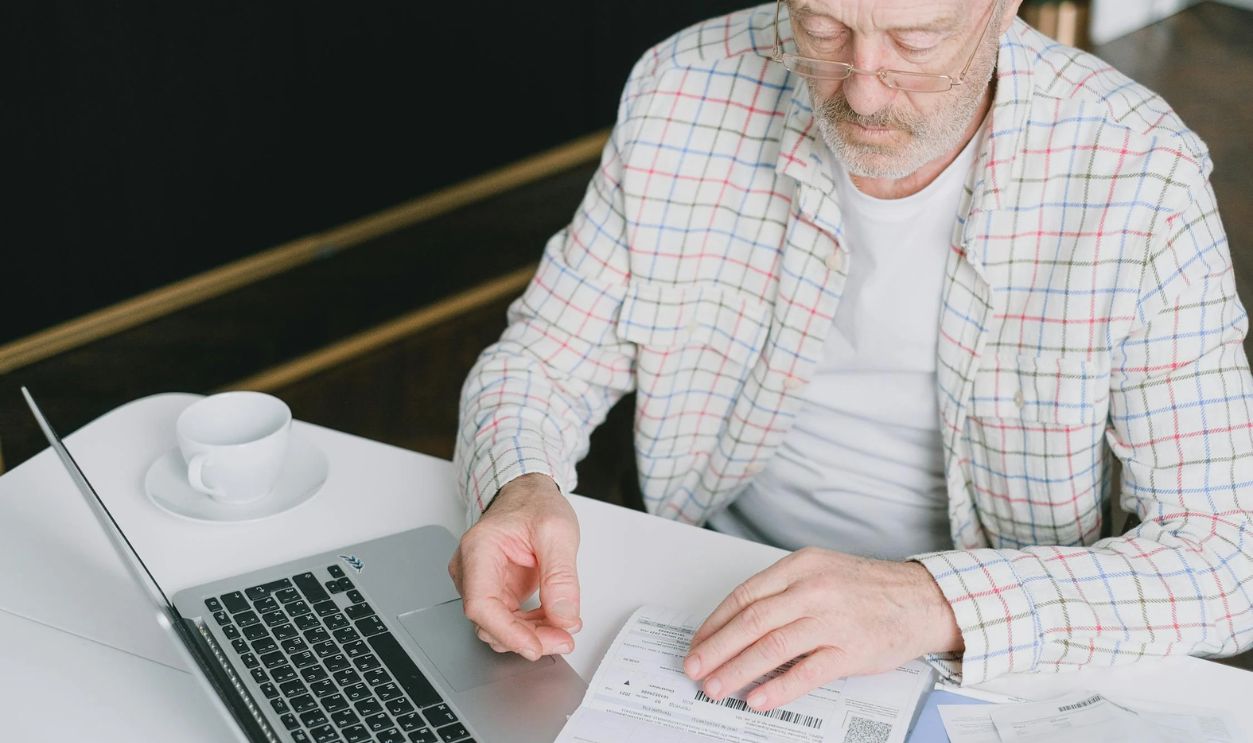 An Elderly Man Looking at the Documents on the Table Near His Laptop