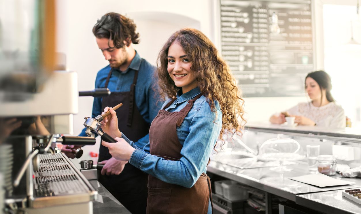 Two young baristas working in coffee shop, standing by counter. University students working part-time in a cafe.