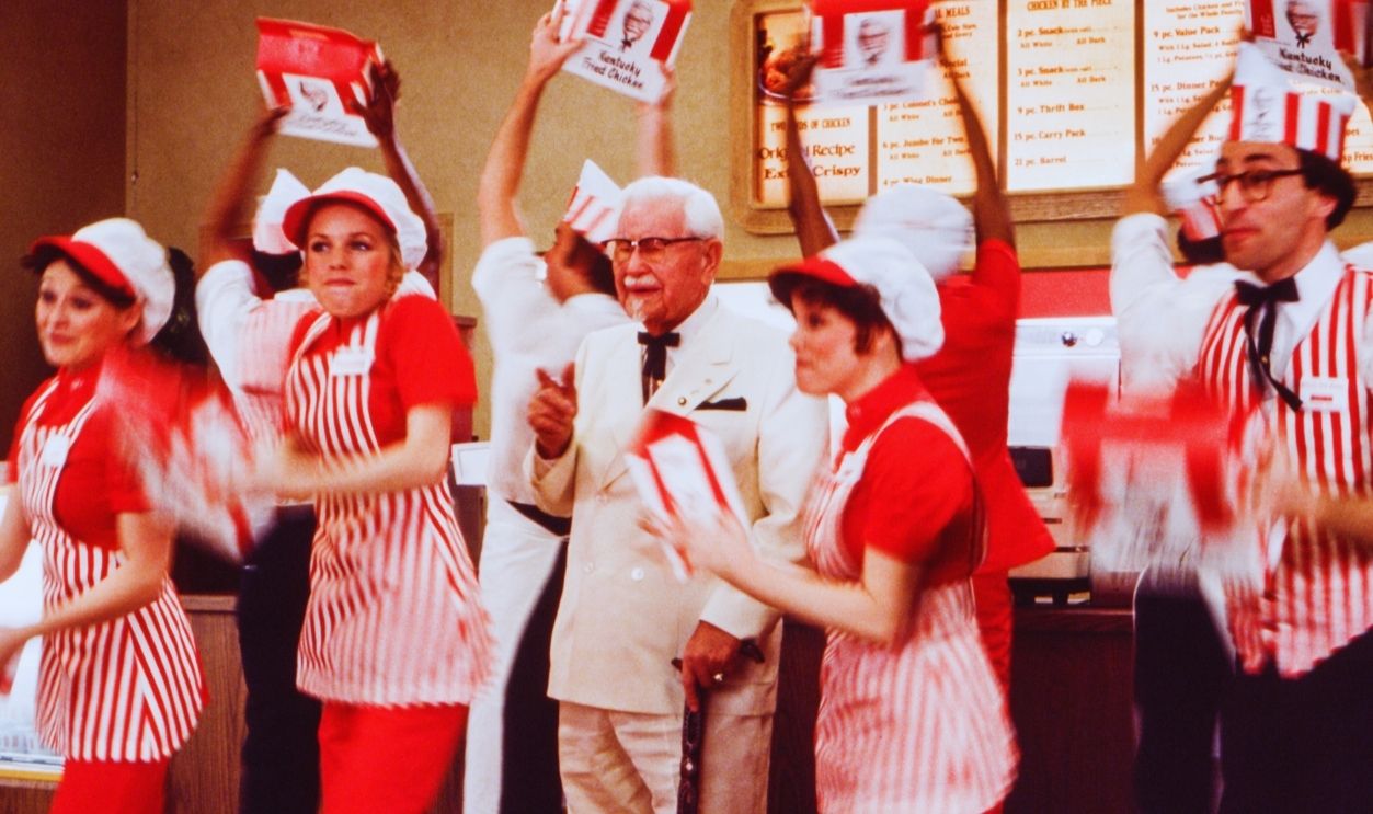 Gettyimages - 1650882834, Kentucky Fried Chicken Commercial Colonel Harland David Sanders (center) who founded the fast food restaurant chain Kentucky Fried Chicken and who is wearing a white suit, a Kentucky Colonel bow tie, lapel pins, a pocket square and eyeglasses is surrounded by actors dressed as employees on a Kentucky Fried Chicken commercial set in a Hollywood studio in February, 1978 in Los Angeles, California.