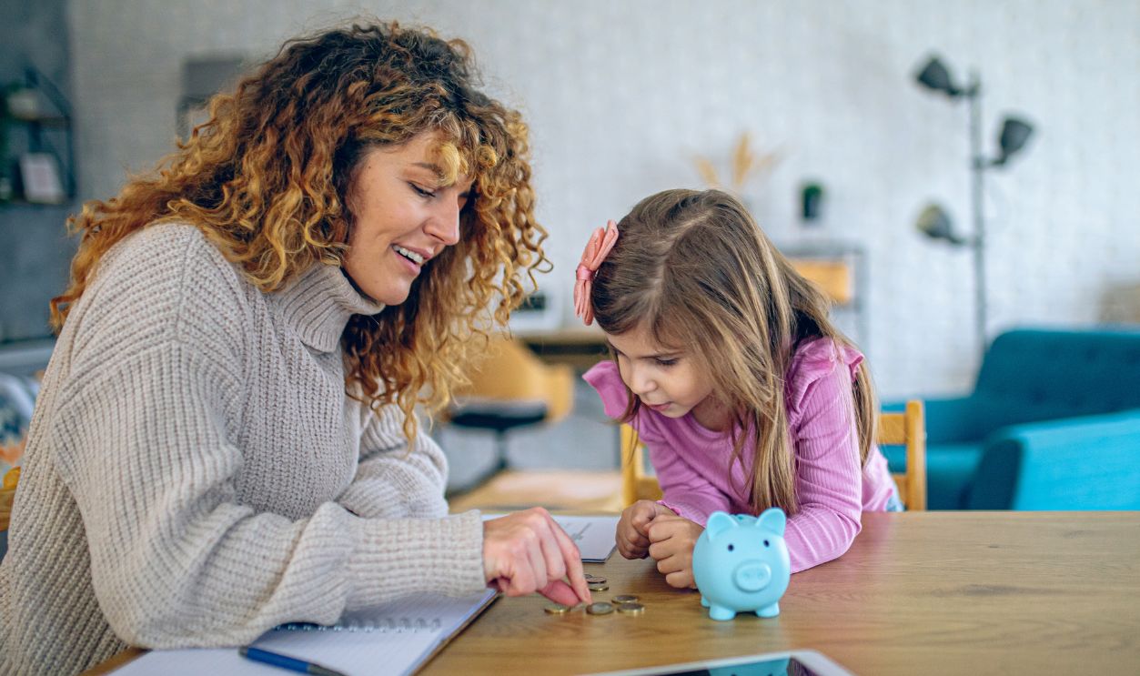Happy mother and her cute daughter putting coin into piggybank, saving money at home