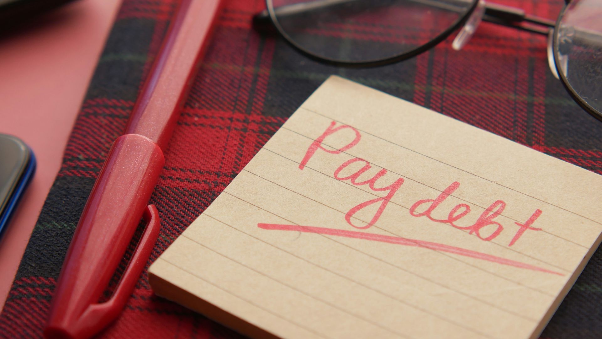 a note that says pay debt next to a pen and glasses