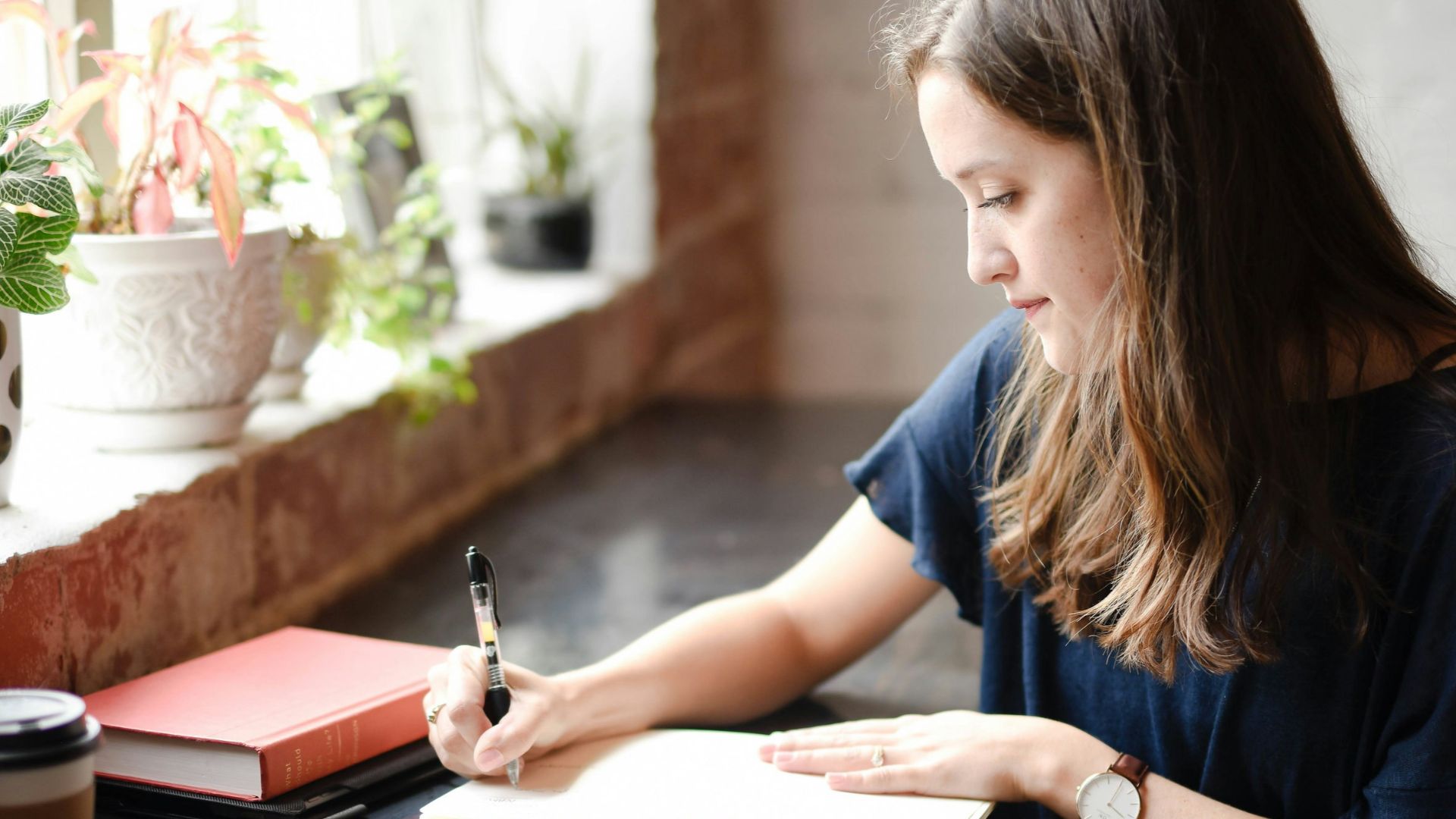 woman sitting in front of black table writing on white book near window