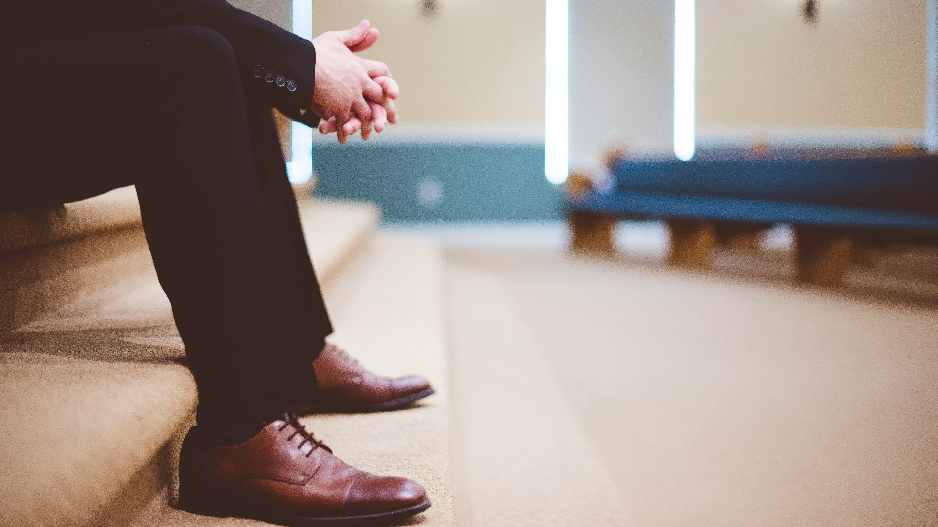 man in black pants and pair of brown leather lace-up shoes sitting on brown carpeted stairs inside room