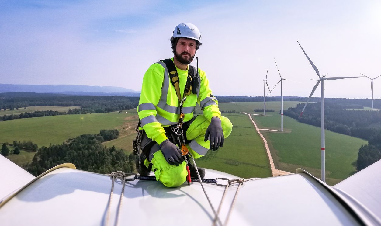 Gettyimages - 1297308942, Industrial climber, rope access technician kneeling on enormous onshore wind-turbine while preparing for blade inspection with wind farm behind him - stock photo Industrial climber, rope access technician kneeling on enormous onshore wind-turbine while preparing for blade inspection with wind farm behind him