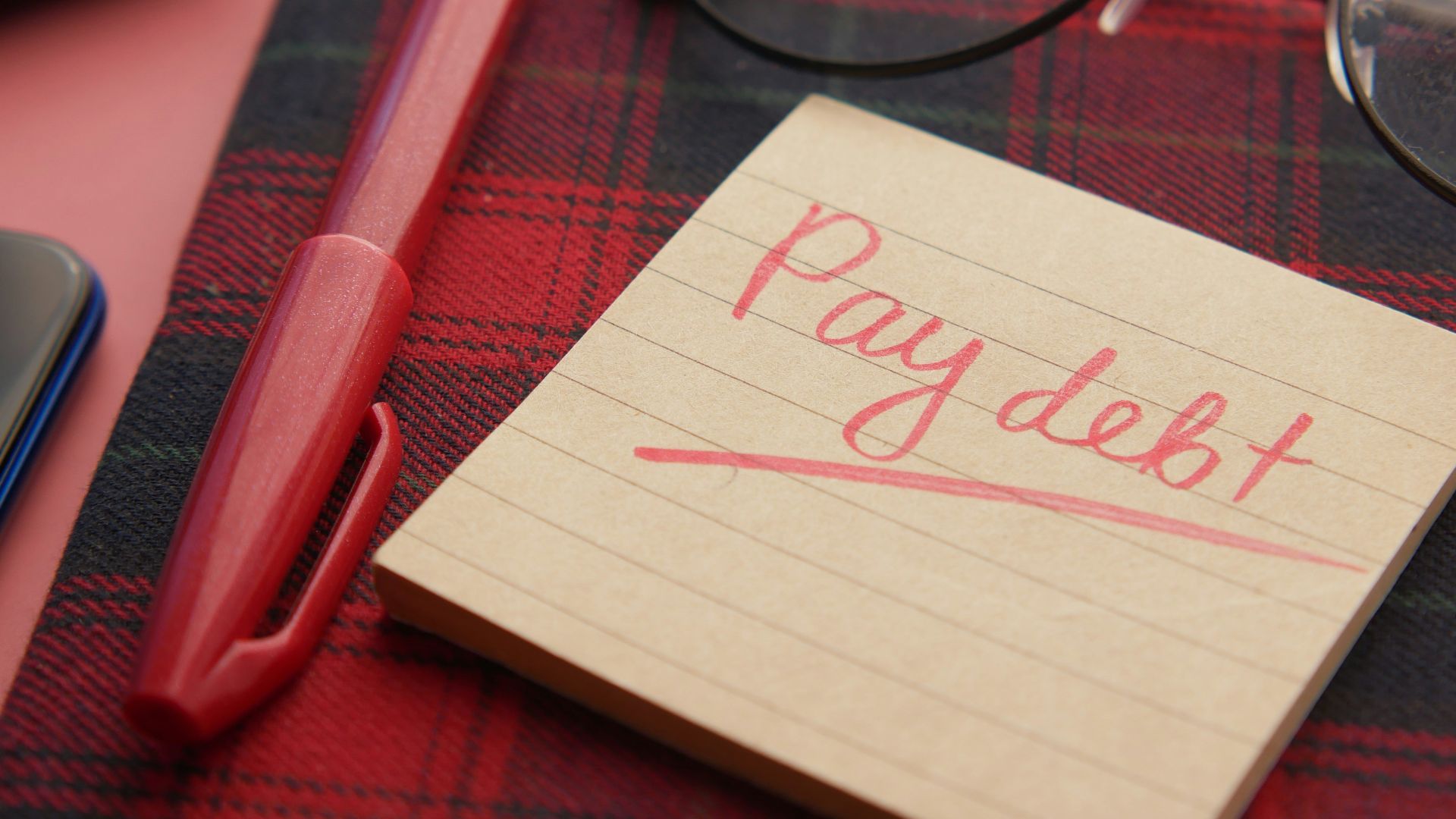 a note that says pay debt next to a pen and glasses