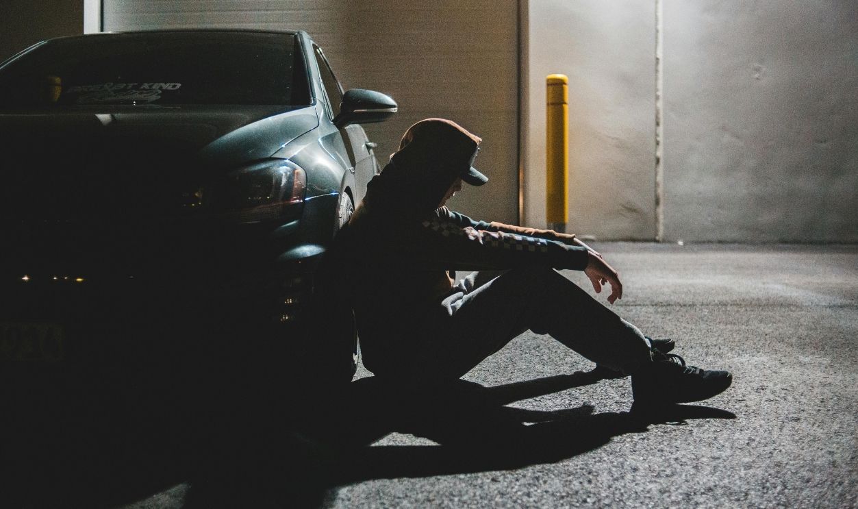 Anonymous upset young guy sitting on ground leaning on car tire