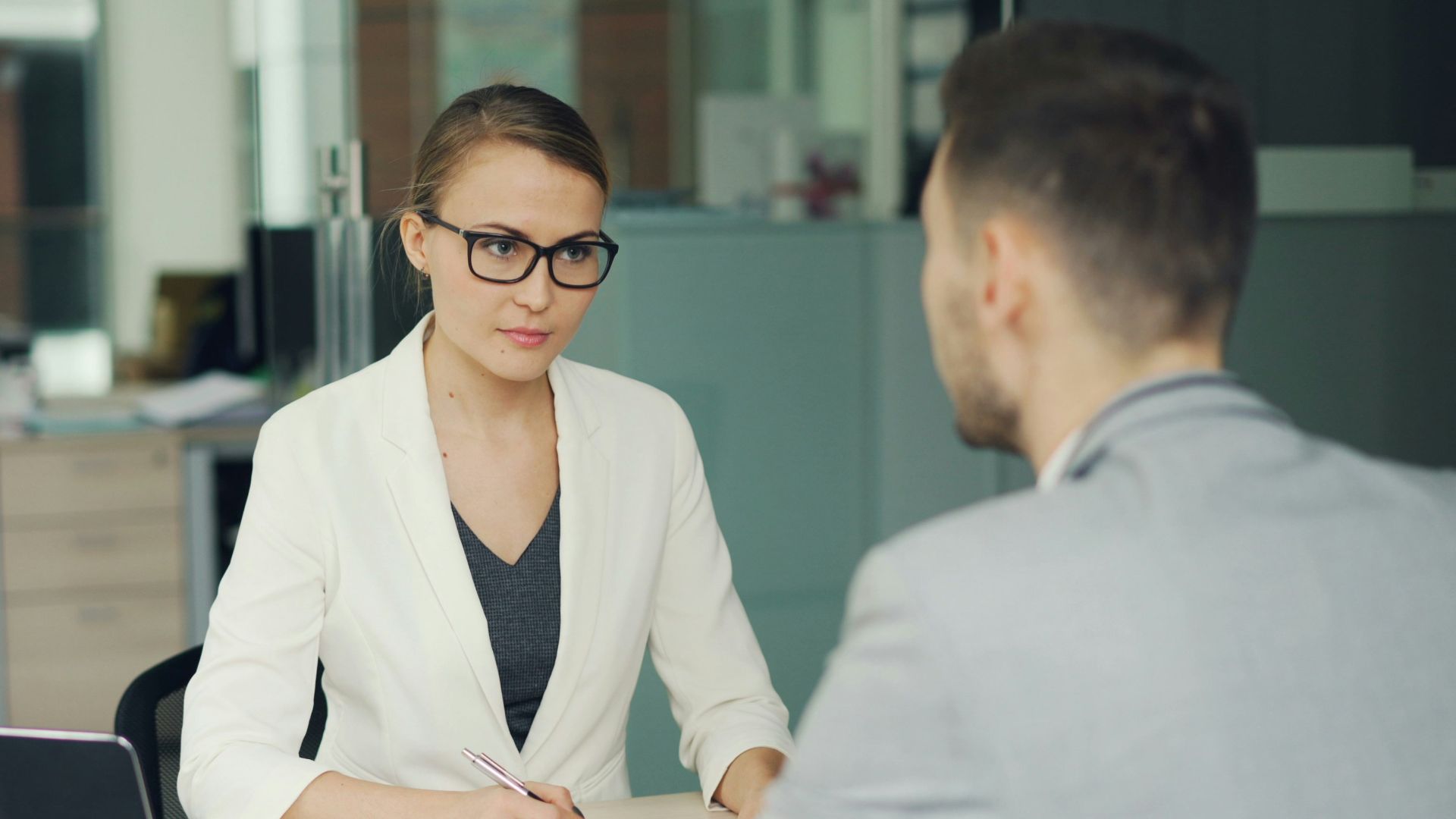 Woman in glasses interviews man at office desk.