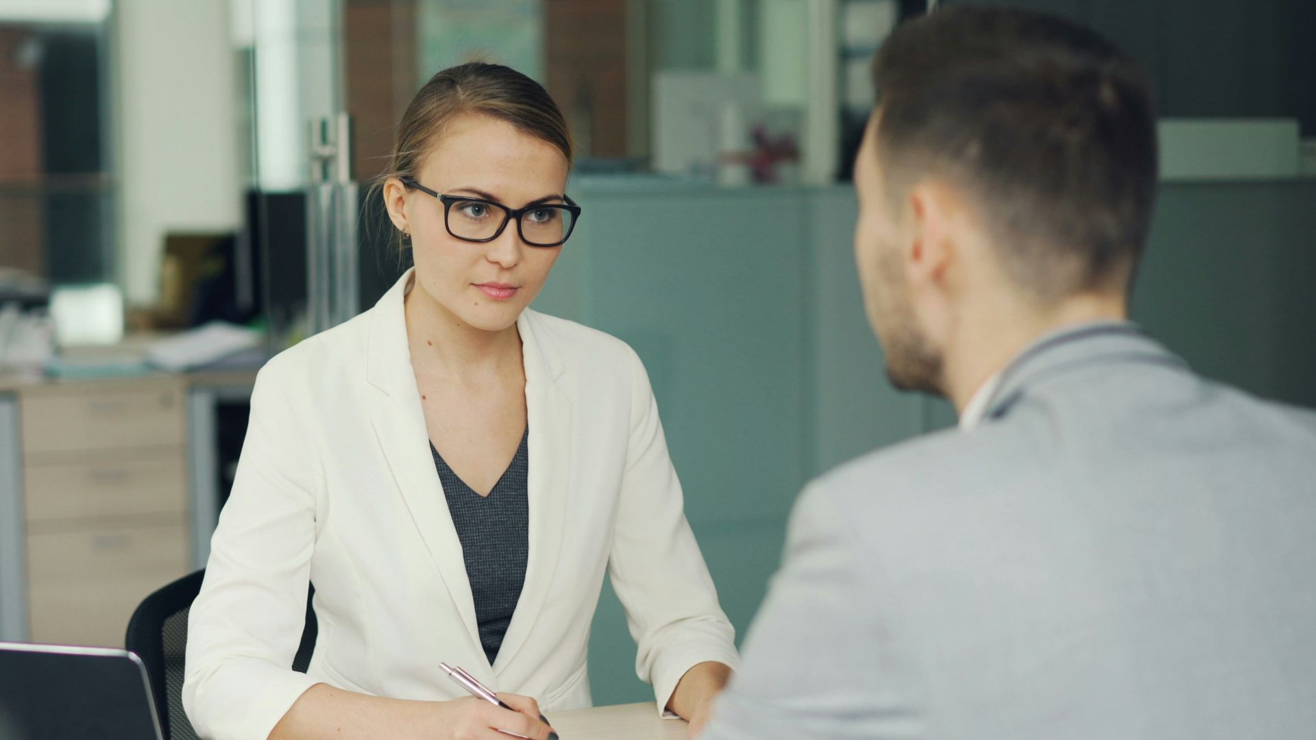 Woman in glasses interviews man at office desk.