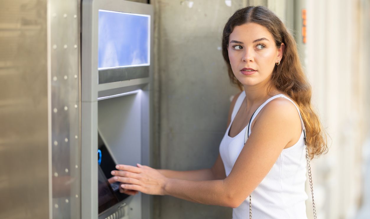 Young girl looking alert and slightly tense while standing at outdoor city ATM, shielding keypad with hand while entering PIN and watching surroundings with caution