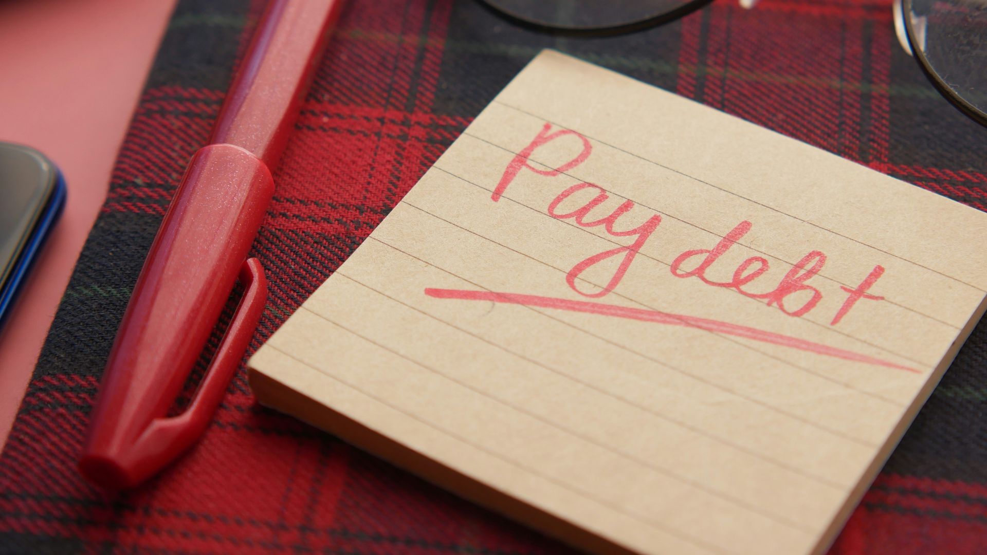 a note that says pay debt next to a pen and glasses