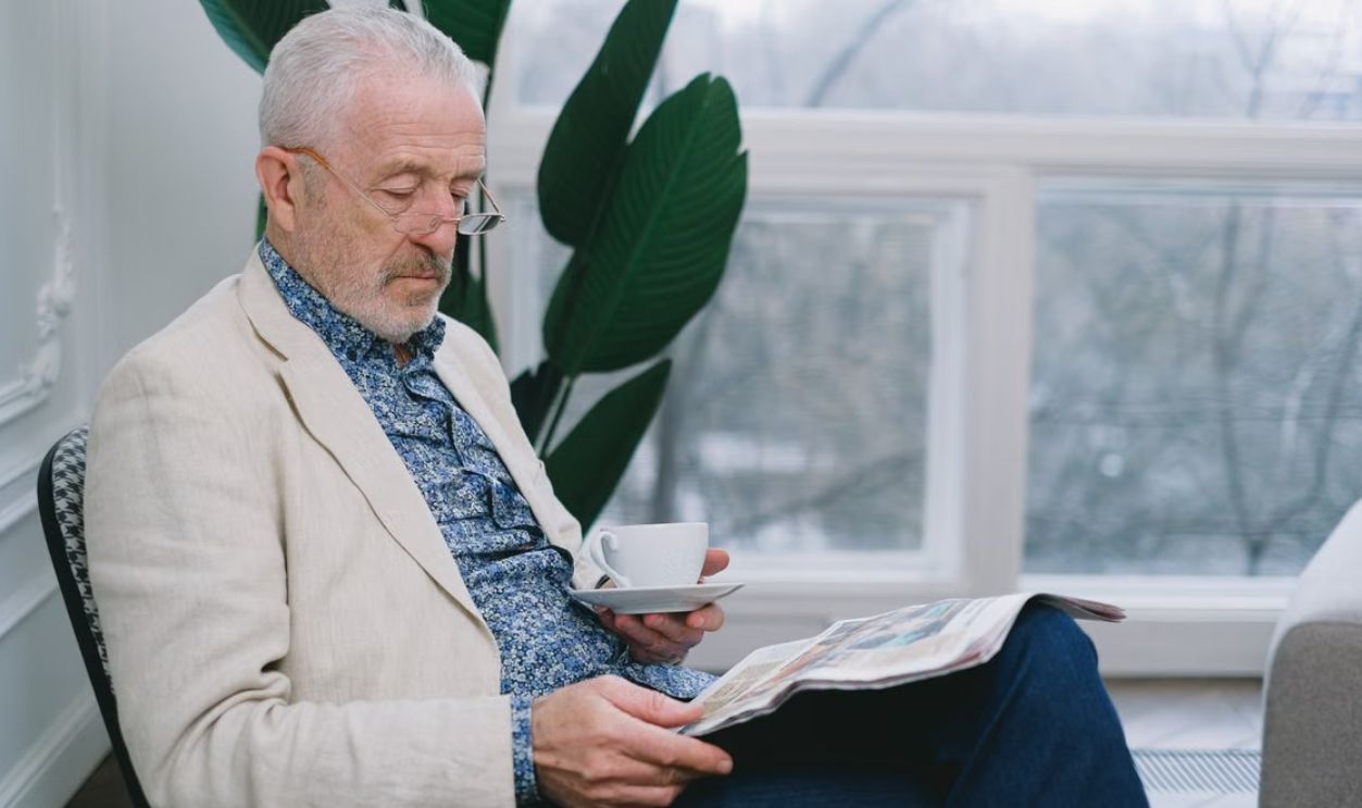 Elderly man sitting indoors, enjoying coffee and reading a newspaper.