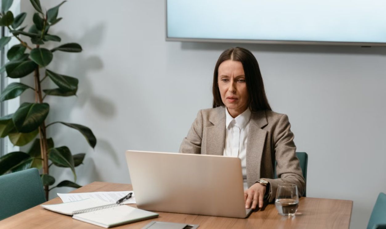 A woman in a business suit using a laptop at an office desk with documents and an indoor plant.