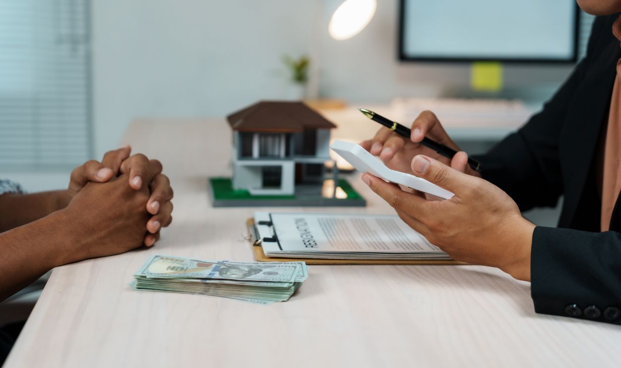 Real estate agent using calculator to calculate house price for client, with house model, contract and money on the table