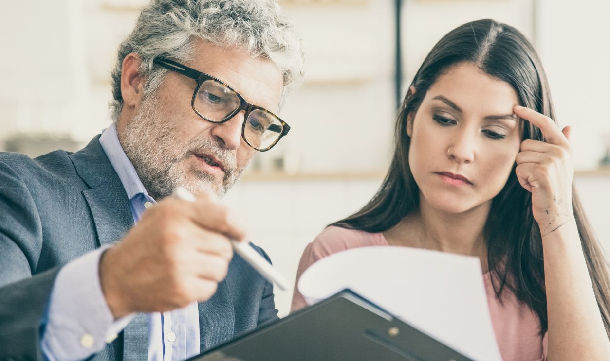 Serious mature legal advisor reading, analyzing and explaining document to female customer. Man and woman sitting at table and reading papers. Expertise and paperwork concept