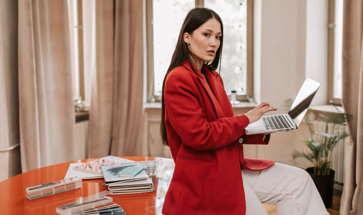 Businesswoman in a red blazer using a laptop at a modern office with creative materials.