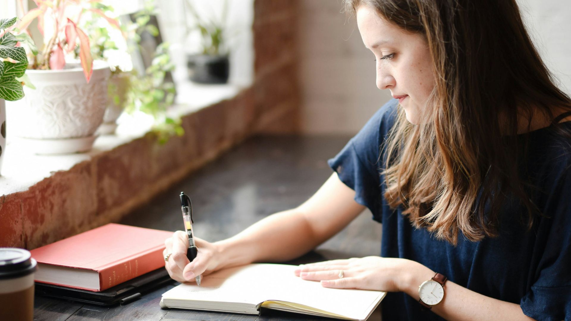 woman sitting in front of black table writing on white book near window